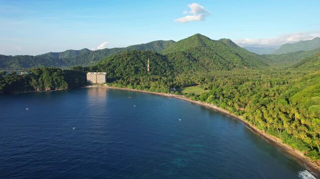 Aerial drone footage of Malimbu beach bay, in Pemenang area, Lombok island, Indonesia, with flat quiet blue water, forests and mountains behind, a building, at golden sunset