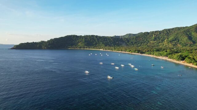 Aerial drone footage of Nipah beach bay, in Pemenang area, Lombok island, Indonesia, with flat quiet blue water, forests and mountains behind, many wooden fishing boats, at golden sunset