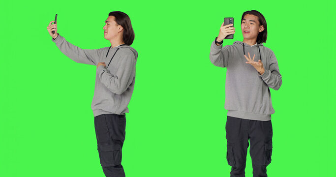 Asian man on videocall connection with friends, using smartphone to call people using phone webcam against green screen, male model talking to someone on videoconference network.