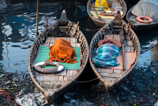 Dhaka Division, Bangladesh - 23 January 2026: View of people sleeping in wooden boats on the polluted banks of the Buriganga River with lifebuoys and scattered trash.