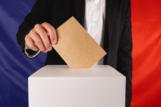 Un homme en costard en train d'ins&eacute;rer une enveloppe dans une urne pour les &eacute;lections en France. En arri&egrave;re plan, le drapeau fran&ccedil;ais.