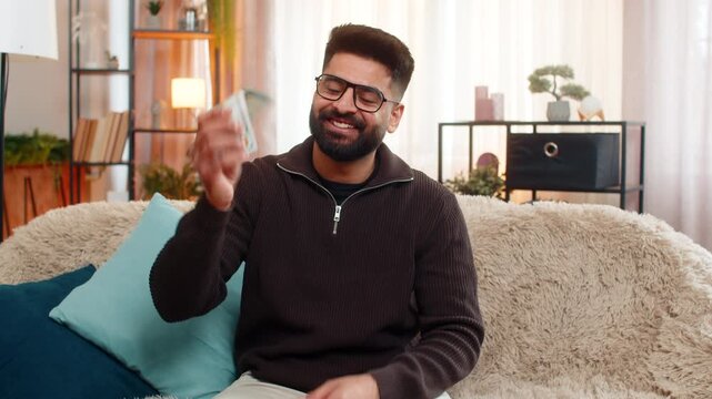 Indian man at home happily counts cash dollars while sitting on sofa, enjoying recent income or lottery win. Hispanic guy expresses joy and financial confidence, celebrating success with satisfaction.