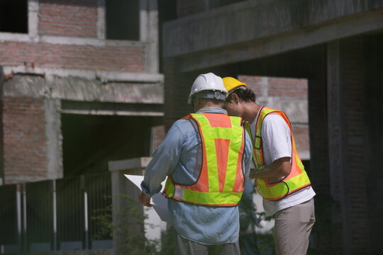 Engineers in hard hats and safety vests review building plans at a residential housing construction site outdoors.
