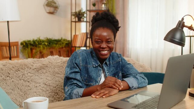 African American young woman sitting at table on home sofa blows a playful air kiss into the camera, smiling with charm and flirty mood. Black girl looks cheerful, expressive, and positive on screen.