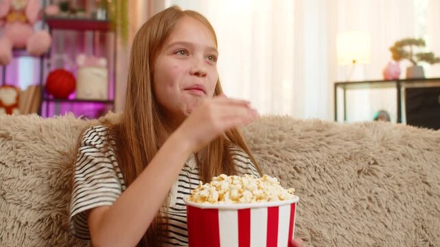 Young child girl eating popcorn and watches funny cartoon on television while sitting on sofa, laughing joyfully, enjoying the show. School kid looks amused and relaxed during cozy movie time at home