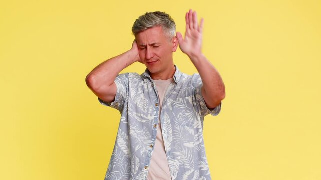 Middle-aged man covering ears with hands and shaking head in clear rejection, refusing to hear or accept something unpleasant. Young guy expressing protest and discomfort isolated on yellow background