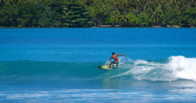 Skilled male surfer makes a top turn on a glassy blue wave framed by dense tropical greenery in Indonesia. Calm, glassy conditions and stunning nature in remote surfing paradise of Mentawai Islands.