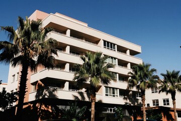Fototapeta premium Modern apartment block with balconies against sunny blue sky. Palm trees frame the facade of the beige building with many windows. Residential architecture.