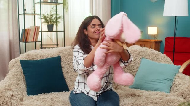 Indian woman at home holds pink bunny toy smiling nostalgic remembering childhood missing child. Arabian girl on sofa gazes at plush rabbit soft toy showing tender warm sentiment longing memory love.