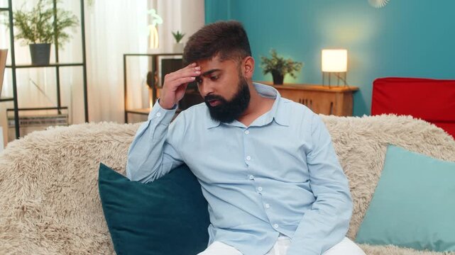 Indian man at home slaps forehead facepalm after smartphone error looks disappointed regretful frustrated. Arabian guy on sofa lowers head grimaces shows guilt mistake reaction stress negative mood.