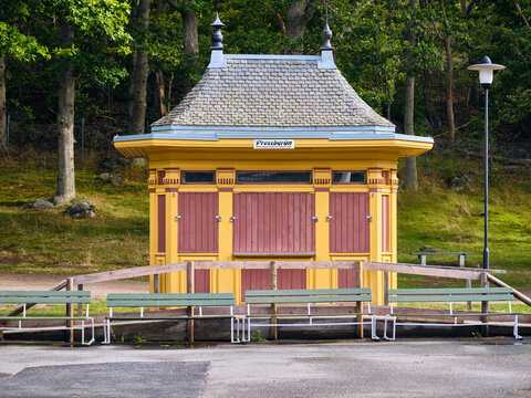 Karlskrona, Sweden - 16 September 2025: View of the historic Pressbyran kiosk in Womoparken with its yellow facade and tiled roof surrounded by green benches and lush trees.