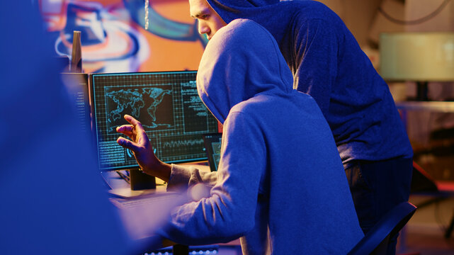 Hacking group founder initiating new member, showing him how to destroy firewalls and infect victim computer in hideout, evil cybersecurity guru teaches script kiddie how to write malware code.