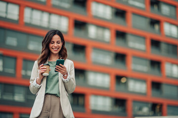 Businesswoman standing outdoors, smiling and checking messages on smartphone while holding a coffee...