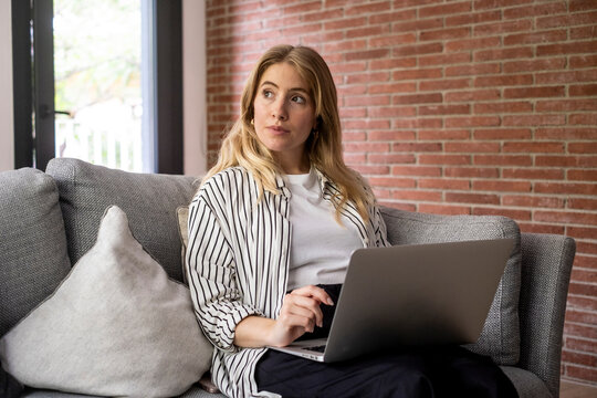 Young woman working on a laptop at home on a sofa, looking thoughtful while using a computer in a modern living room with exposed brick wall and natural light