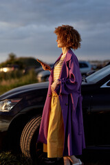 Fototapeta premium A stylish woman stands beside a dark car during golden hour, wearing a purple trench, striped top, and yellow dress. A candid lifestyle moment about confidence, freedom, and urban exploration.