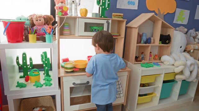 Boy in blue shirt pouring a toy cup into a play sink while raising his hand at a children's play kitchen in building; imaginative curiosity.