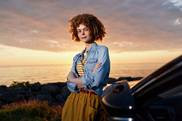 Fototapeta premium Confident woman with curly hair leaning against a car at sunset, wearing a denim shirt and striped top. Lifestyle portrait showcasing outdoor style, independence, and warm, relaxed mood.