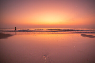 Lone figure stands in the ocean waves during a vibrant sunset with a crescent moon © narongcp