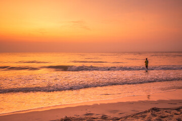 Fototapeta premium Lone figure wading into the ocean at sunrise with warm golden light reflecting on the water