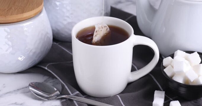 Making tea. Woman putting teabag into cup with hot water and adding sugar at white marble table, closeup