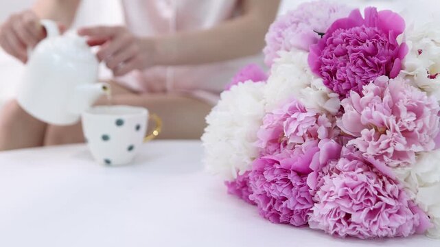 Woman pouring tea into cup and bouquet of beautiful peony flowers on bed indoors, selective focus. Camera moving in