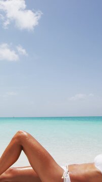 Vertical video of Beach body woman sun bathing on travel vacation. Sexy bikini body woman relaxing sun tanning lying down on perfect paradise beach at tropical luxury destination in the Caribbean.