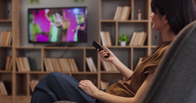 Woman watching television with remote on chair. She enjoys relaxation at home in a living room, with entertainment and digital media supporting a relaxed lifestyle. Clear concept of home media.