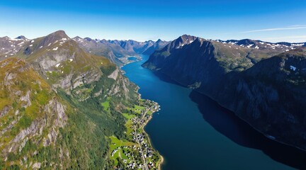 Fototapeta premium Aerial view of deep blue fjord between tall green mountains. Small village rests on green shore beside calm water. Bright clear blue sky covers peaceful natural landscape.