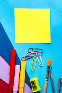 Blank yellow sticky note on colorful blue background with markers paperclips paintbrush compass and sharpener back to school copy space. 