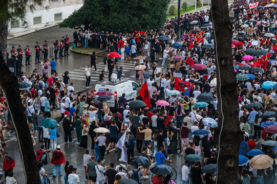 Tirane, Albania - 03 June 2026: View of a large crowd of protesters gathered on a city street with Albanian flags and umbrellas during a demonstration near a news van and police line.