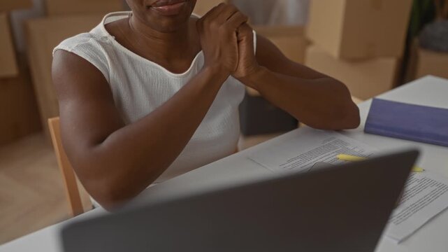 Woman at laptop with clasped hands and documents on table, moving boxes stacked nearby in a building; calm focus.
