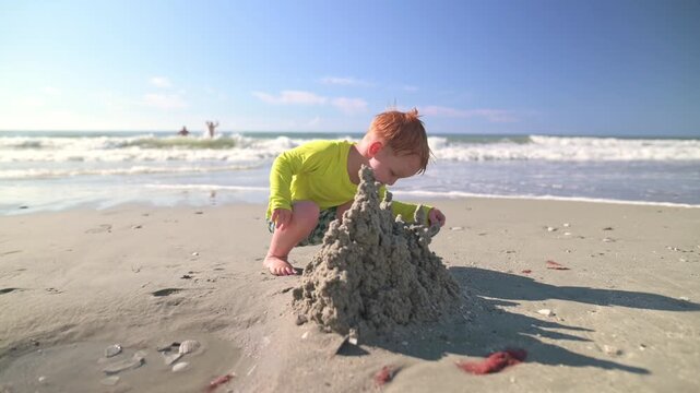 Young boy in a bright yellow shirt kneels on the sandy beach, carefully shaping a sandcastle as waves roll in, with distant figures enjoying the sun and surf under a clear blue sky