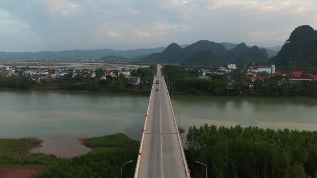 Bridge Crossing Calm River Into Small Town, Vehicles Moving Toward Clustered Houses With Karst Hills Beyond, Soft Dusk Glow Over Water And Settlement In Phong Nha Vietnam