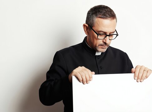 Mature priest in clerical collar and black cassock holds blank white sign. Man with glasses looks down thoughtfully, holding empty board for message. Male clergy offers space for text.