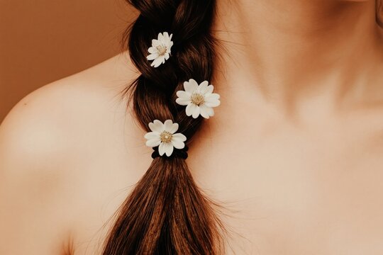 Woman dark braided hair adorned with white flowers. Soft brown backdrop and skin tones create serene, natural beauty. Minimalist, dreamcore aesthetic for wellness and fashion.