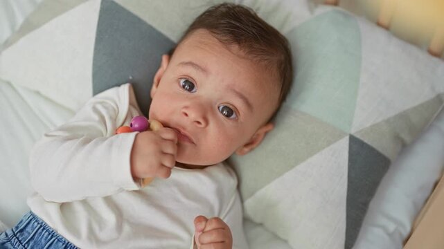 Baby boy holding wooden teether to mouth, sucking with hand visible while lying in crib bed on patterned pillow; quiet curiosity.