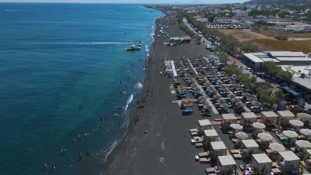 Aerial View of Perissa Black Sand Beach Resort and Coastal Promenade in Santorini Greece. A long stretch of dark volcanic black sand lined with organized rows of thatched umbrellas.