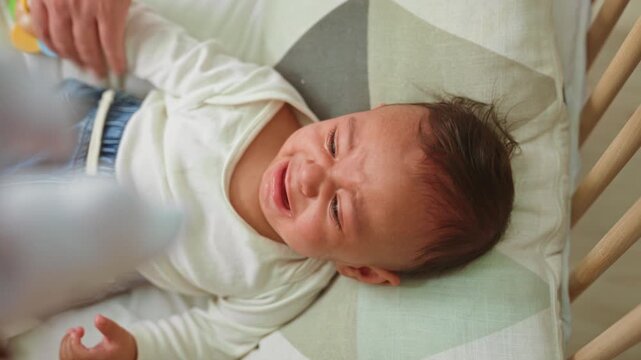 Boy clutching colorful wooden rattle with hand, face scrunched and crying on cradle bed in building; need comfort distress.