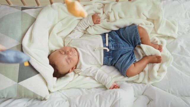 Hispanic baby boy sleeping on crib blanket with bare feet and clenched fist in nursery; calm peaceful rest.
