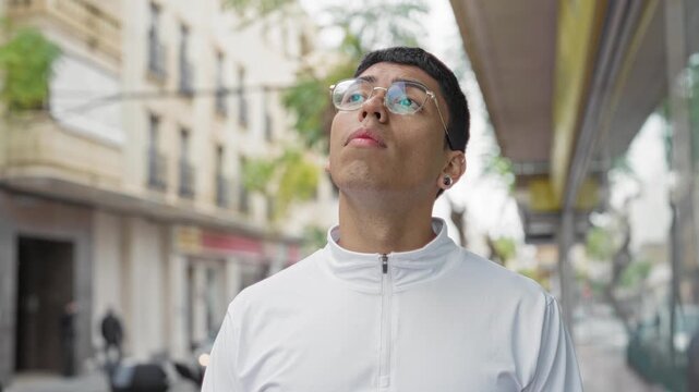 Man wearing glasses and white zip jacket looking up on street near storefront and building facade; curiosity.