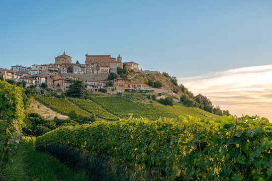 View of the historic hilltop village and lush green vineyards under a soft sunset sky with church towers and stone buildings in La Morra, Piedmont, Italy.
