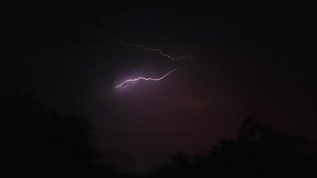 Stunning lightning storm strikes dark night sky above silhouettes of trees capturing dramatic atmospheric electrical display.