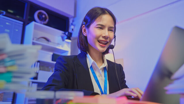 Smiling young Asian customer service representative wearing microphone headset and working on laptop in modern office.