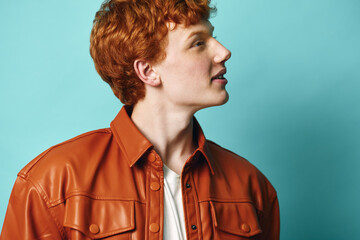 Young man with red hair wearing brown leather jacket and white shirt looking sideways on blue background. Profile portrait with casual style and bright natural color contrast. © SHOTPRIME STUDIO