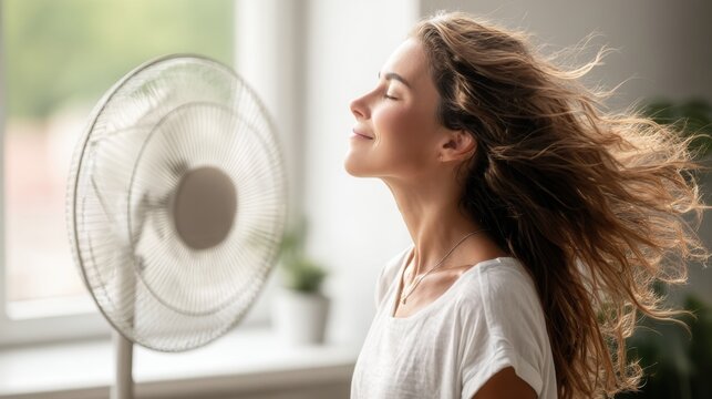 Young woman enjoying refreshing breeze from electric fan during hot summer day at home for cooling