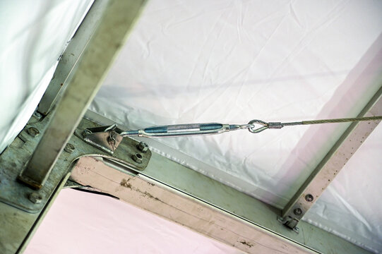 A detailed shot of the indoor metal structure supporting a white tent or canopy ceiling, showing a turnbuckle tensioner connected to a steel wire cable