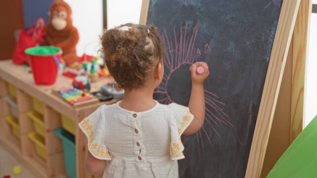 Little girl holds pink chalk and draws a sun on a blackboard, hand and finger visible in a studio; curiosity imagination play.