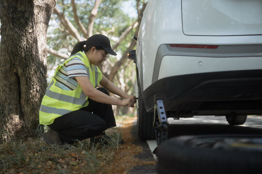 Woman fixes flat tire beside road in daytime near trees