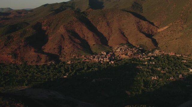 Amazigh mountain village occupies a fertile terrace in Ourika Valley, Al Haouz Province, Marrakesh-Safi, Morocco, beneath rugged High Atlas slopes, aerial tracking shot.