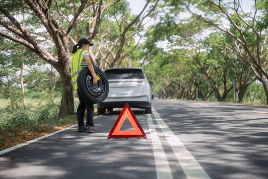 Person holds a spare tire on a road with trees and a car in the background during daytime